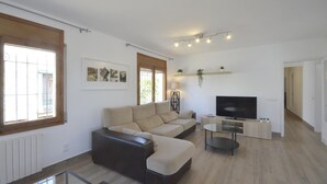 A modern living room with a sofa, a glass coffee table, and a TV. There are windows with blinds, a plant on a shelf, and a radiator. - To rent house with private pool in Begur, center (Begur)