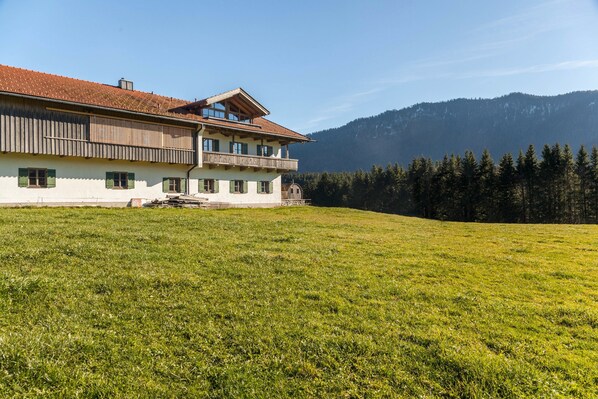 A two-story building with a balcony, a grassy field in the foreground, and a forested mountain in the background.