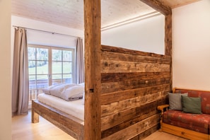 Una cama con estructura de madera, vista a un paisaje verde a través de una ventana, pared de madera y un sofá rojo con cojines estampados.