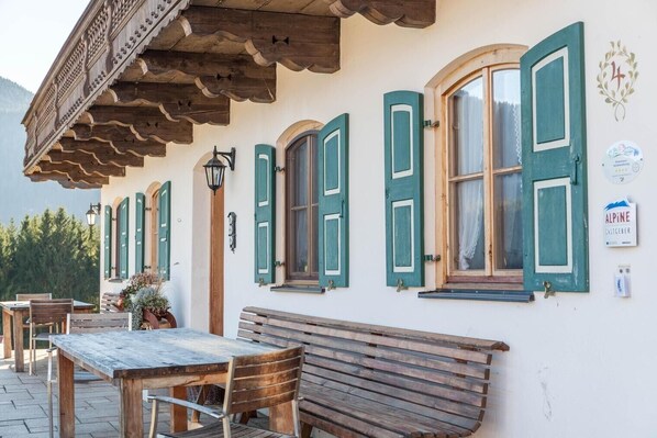 Un edificio tradicional con bancos y mesas de madera, contraventanas verdes y un letrero que indica una ubicación alpina.
