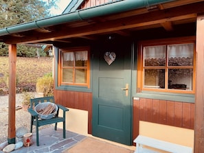 Una cabaña de madera con puerta verde, dos ventanas con cortinas y un banco afuera.