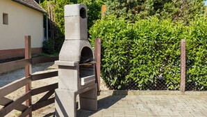 A stone outdoor grill structure with a chimney, surrounded by a wooden fence and a hedge. - Vacation home Tamas by Interhome (Balatonoszod)