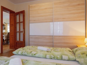 A bedroom with a wooden wardrobe, a bed with floral bedding, and a visible dining area in the background.