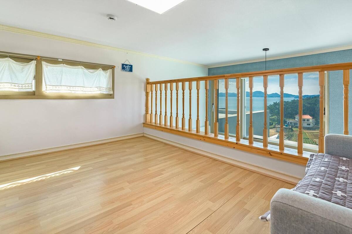 Een ruime kamer met een houten vloer, een grijze bank, een raam met witte gordijnen en uitzicht op water en bergen.
