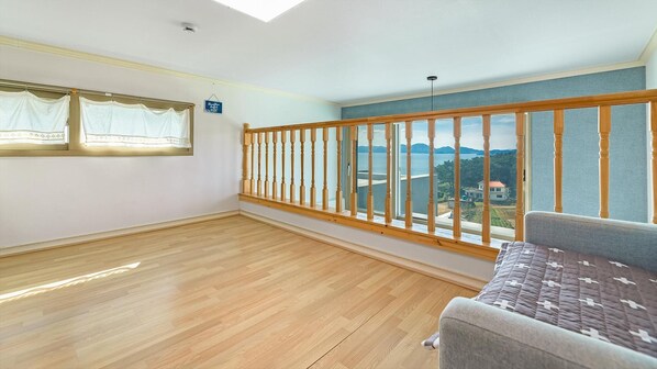 A spacious room with a wooden floor, a grey sofa, a window with white curtains, and a view of a body of water and mountains.