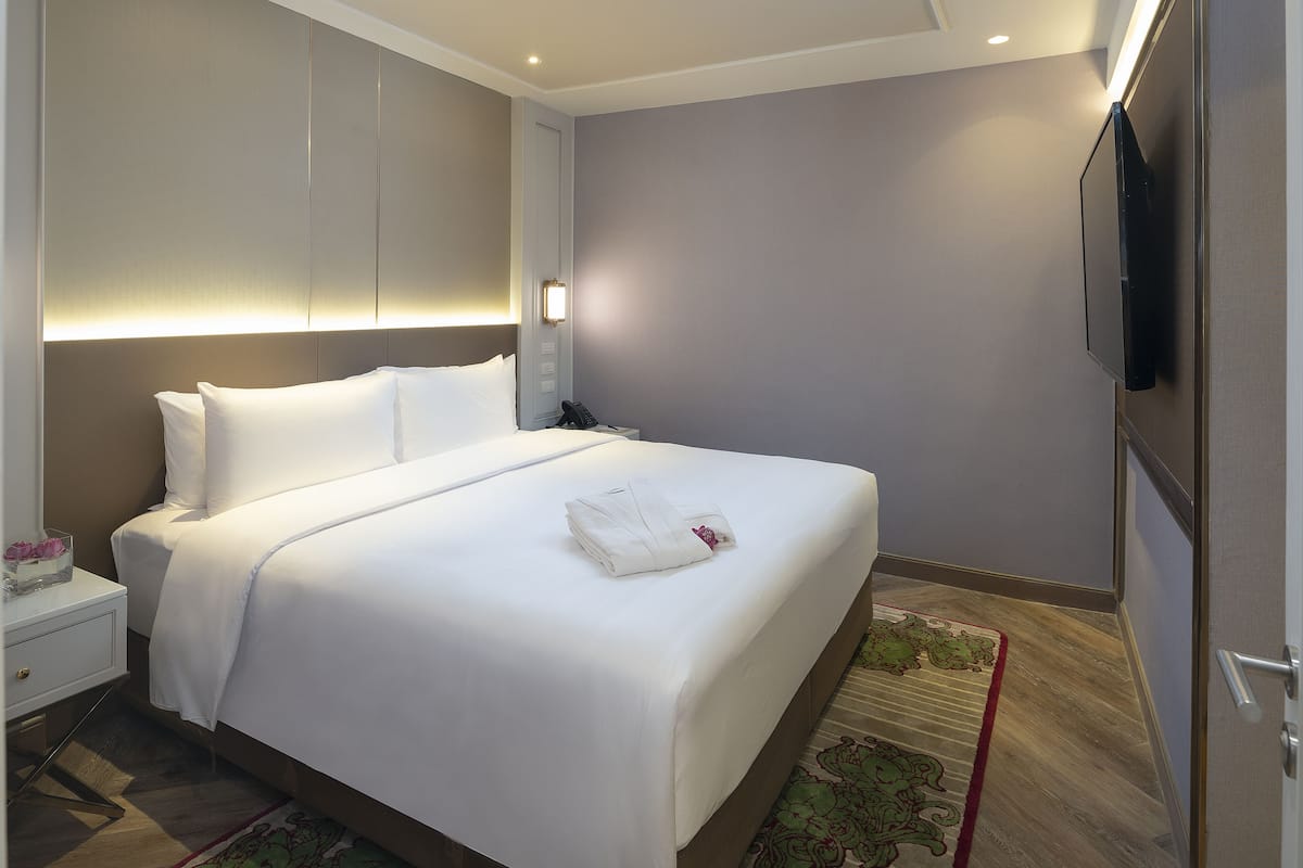 Una cama bien hecha con sábanas blancas y una toalla doblada, una mesita de noche con lámpara, y un televisor montado en la pared.
