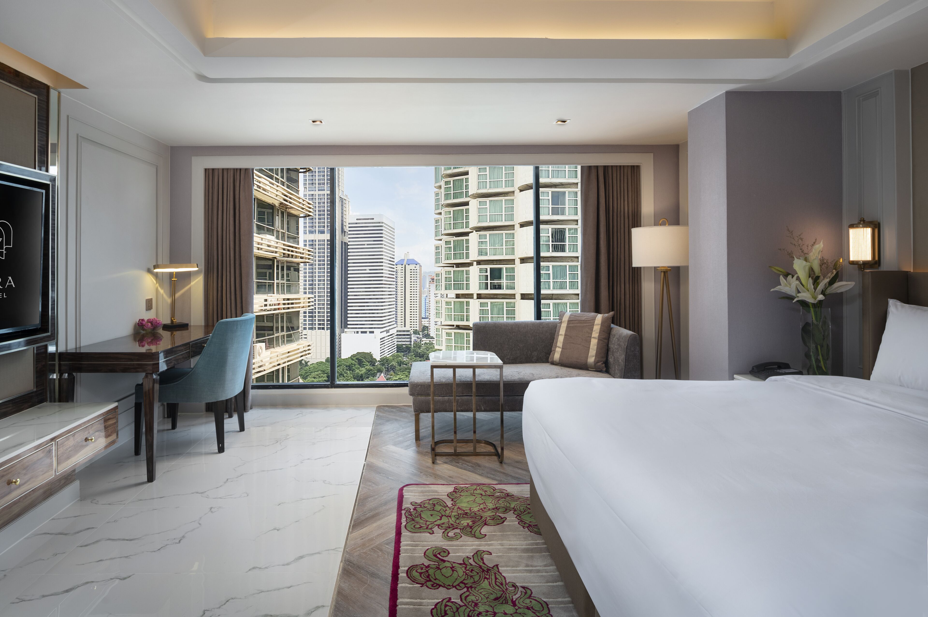 A modern hotel room with a large bed, a desk with a chair, a TV, and a view of the cityscape through large windows.