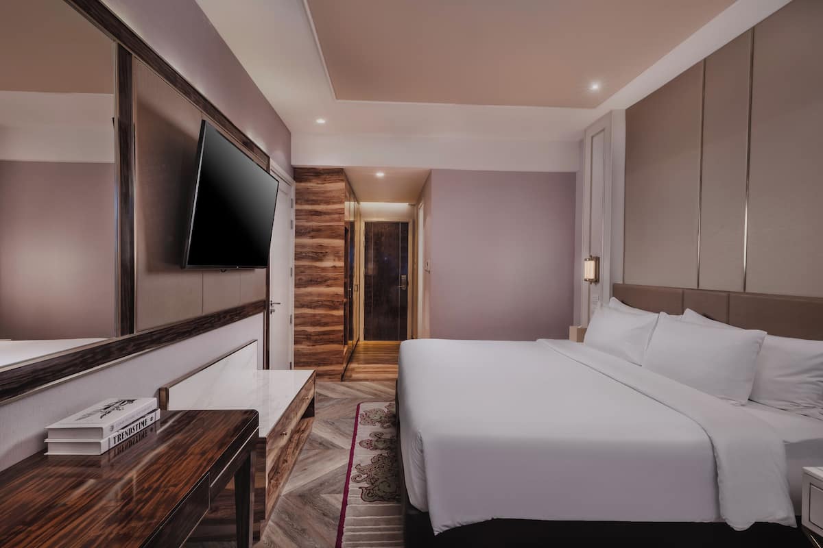 Habitación de hotel con cama, televisor montado en la pared, banca de madera y puerta que da a otra habitación.