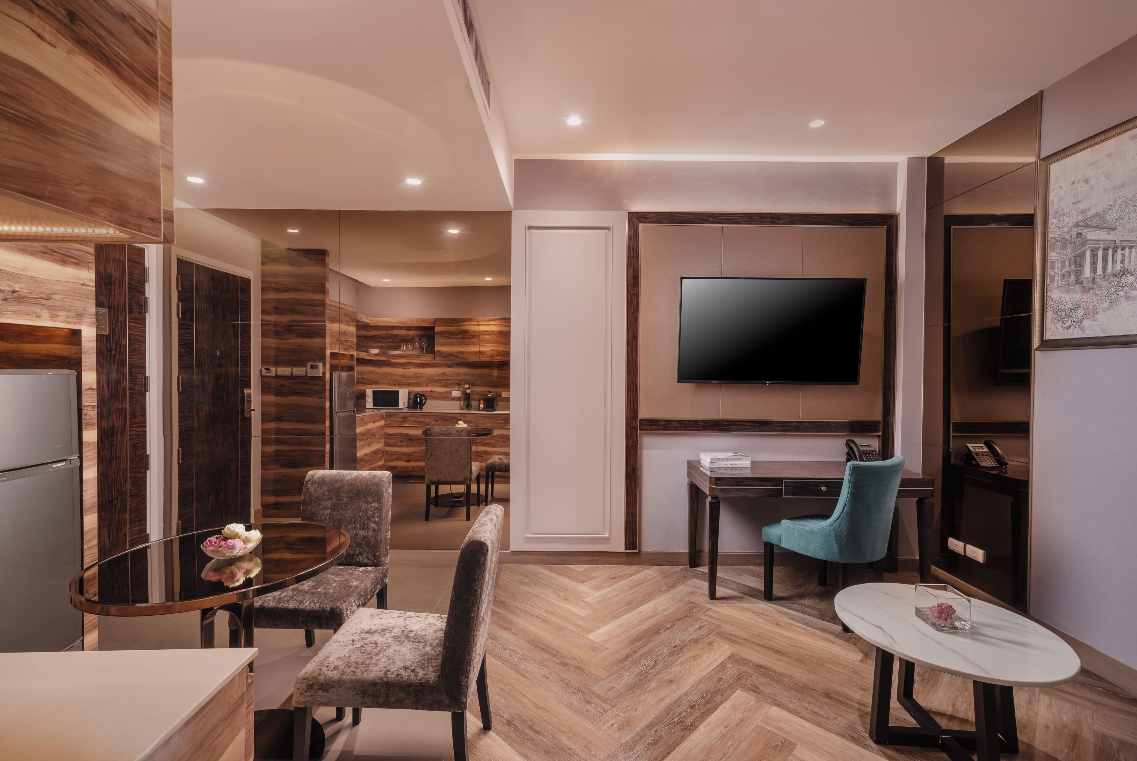 A modern living room with a herringbone wood floor, a flat-screen TV mounted on the wall, a dining area with a glass table and chairs, and a kitchen area in the background.