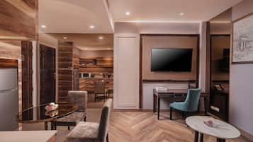 A modern living room with a herringbone wood floor, a flat-screen TV mounted on the wall, a dining area with a glass table and chairs, and a kitchen area in the background.