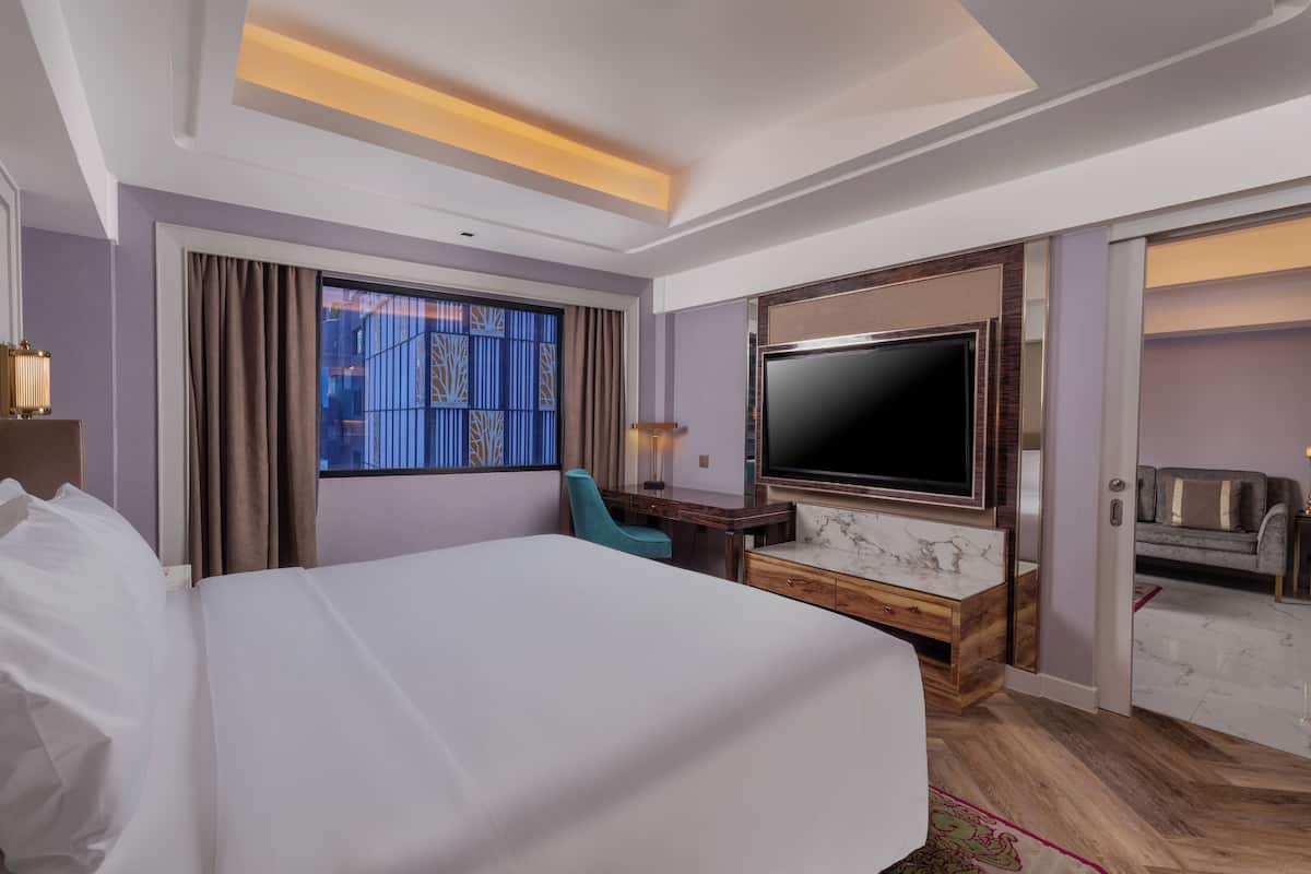 Habitación de hotel con una cama grande, un escritorio con televisor, una silla y vistas a un edificio desde la ventana.