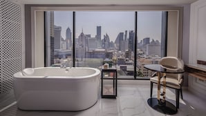 Ein modernes Badezimmer mit einer großen Badewanne, einem kleinen Tisch mit Stuhl und Blick auf die Stadt durch große Fenster.