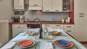 A compact kitchen with white cabinets, a coffee machine, a stove, and a dining table set for four with colorful plates and glasses. - Cottage al Lago by Interhome (Gravedona)