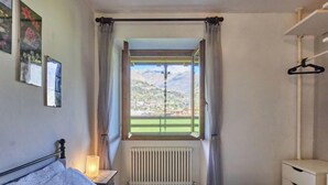 A bedroom with a bed, a bedside table with a lamp, a radiator, a window with curtains, a hanging rack, and a view of a mountainous landscape. - Cottage al Lago by Interhome (Gravedona)