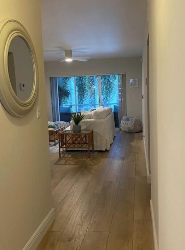 A hallway with a wooden floor, a white sofa, a small table with a plant, and a large window with a view of trees. - 2BR Condo in Gardens by the Sea – 3-Min Walk to Secluded Beach + Heated Pool (Pompano Beach)
