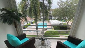 A balcony with wicker furniture and a small table, overlooking a swimming pool and greenery.