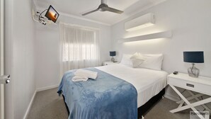 Een moderne hotelkamer met een bed, een plafondventilator, een aan de muur bevestigde televisie en een nachtkastje met een lamp.