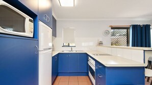 Een compact keuken met blauwe kastjes, een witte magnetron, een spoelbak en een raam met gordijnen.