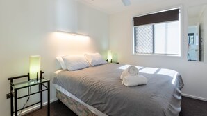 A neatly made bed with a grey blanket, two pillows, and a rolled towel on the side. There is a ceiling fan above the bed, a bedside table with a lamp, and a window with a view of the outside.