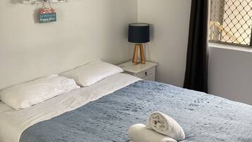A neatly made bed with a blue blanket and white pillows, a bedside lamp, a wall-mounted air conditioner, and a window with a view of a building.