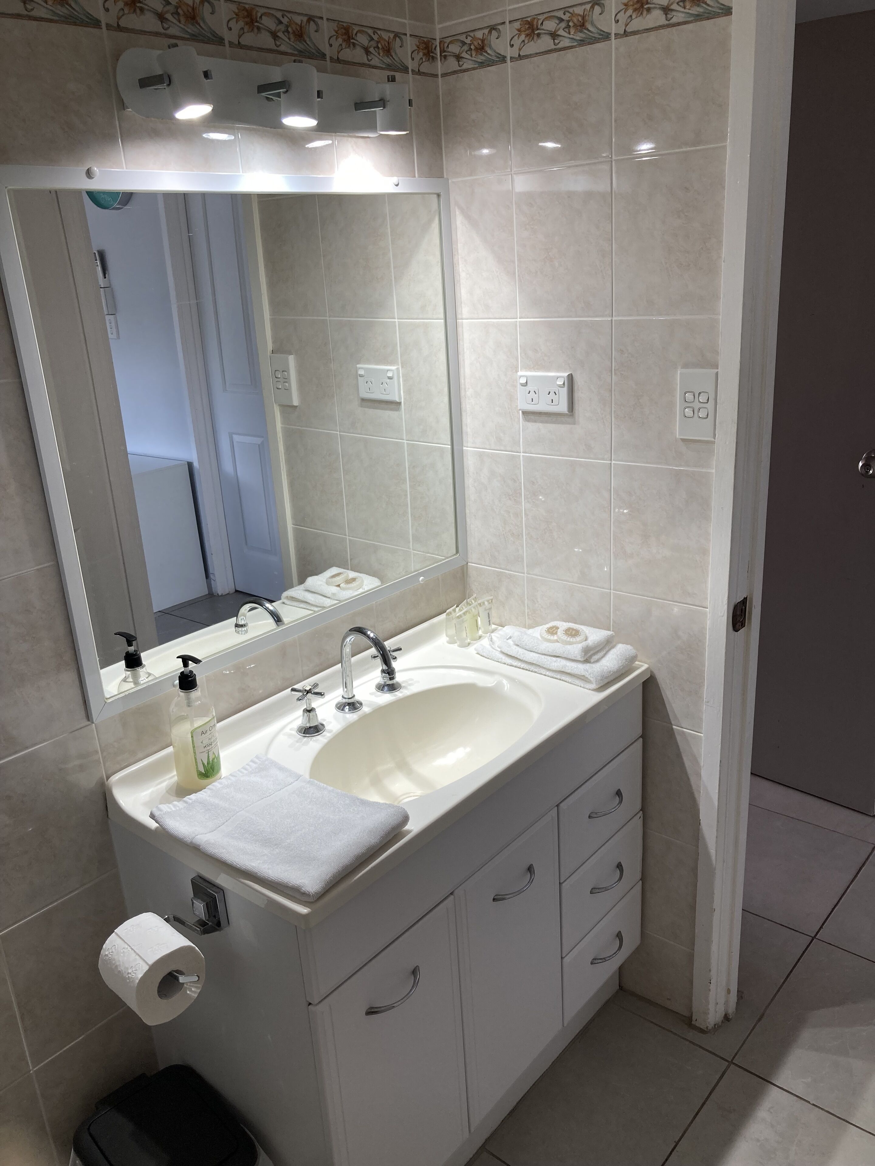Une salle de bain avec un lavabo blanc, un miroir et un meuble-lavabo avec des tiroirs. Il y a une serviette sur le comptoir et un flacon de savon pour les mains.