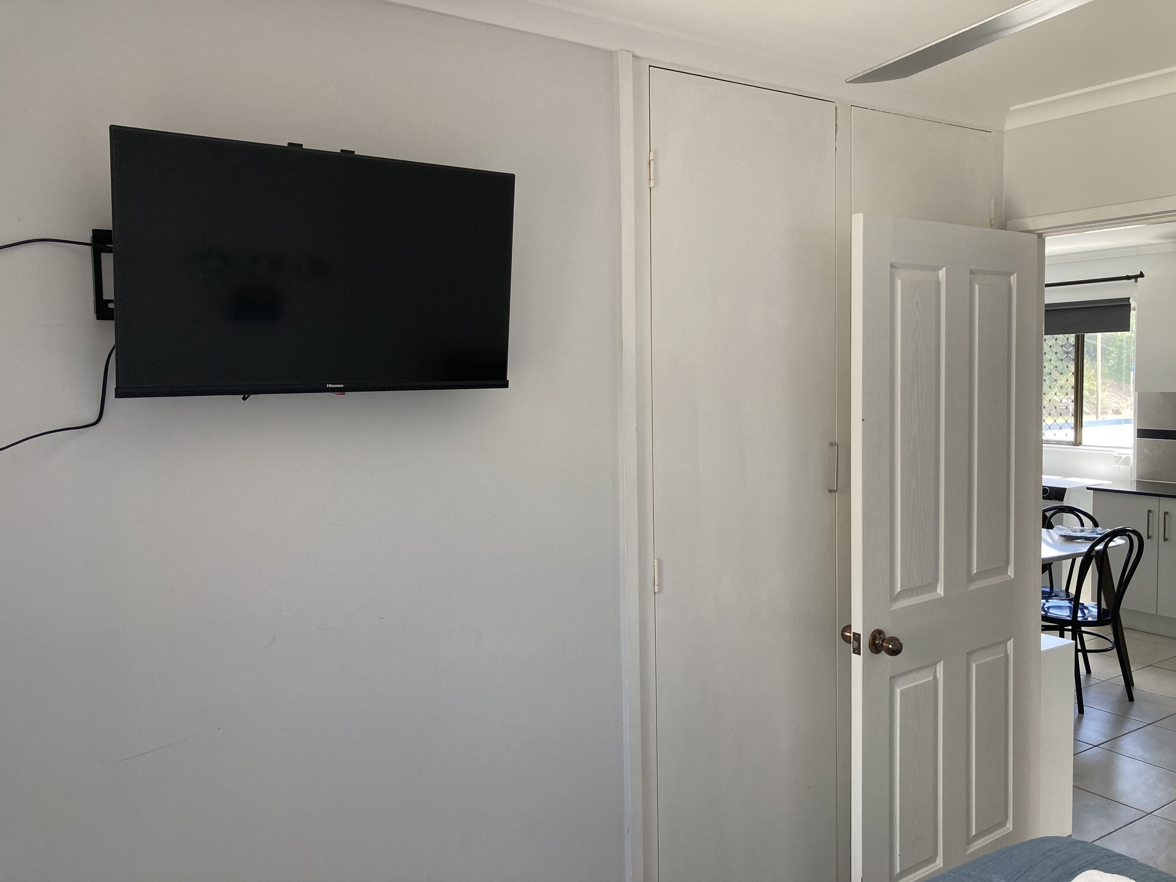 A room with a mounted television on the wall, a white door, and a glimpse of a dining area with a table and chairs.