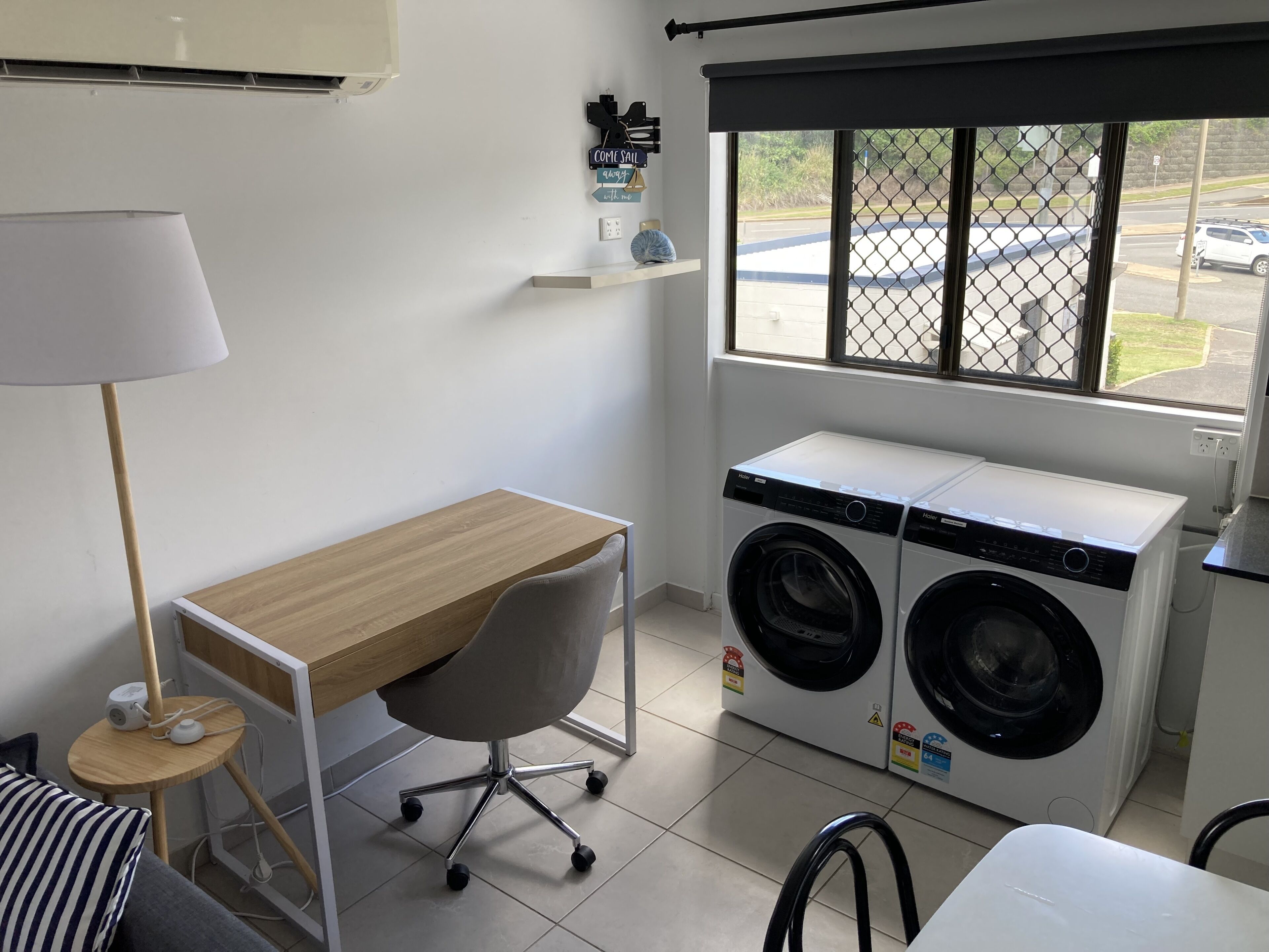 A compact laundry room with a wooden desk, two front-loading washing machines, a chair, and a window with a view of a parking lot.