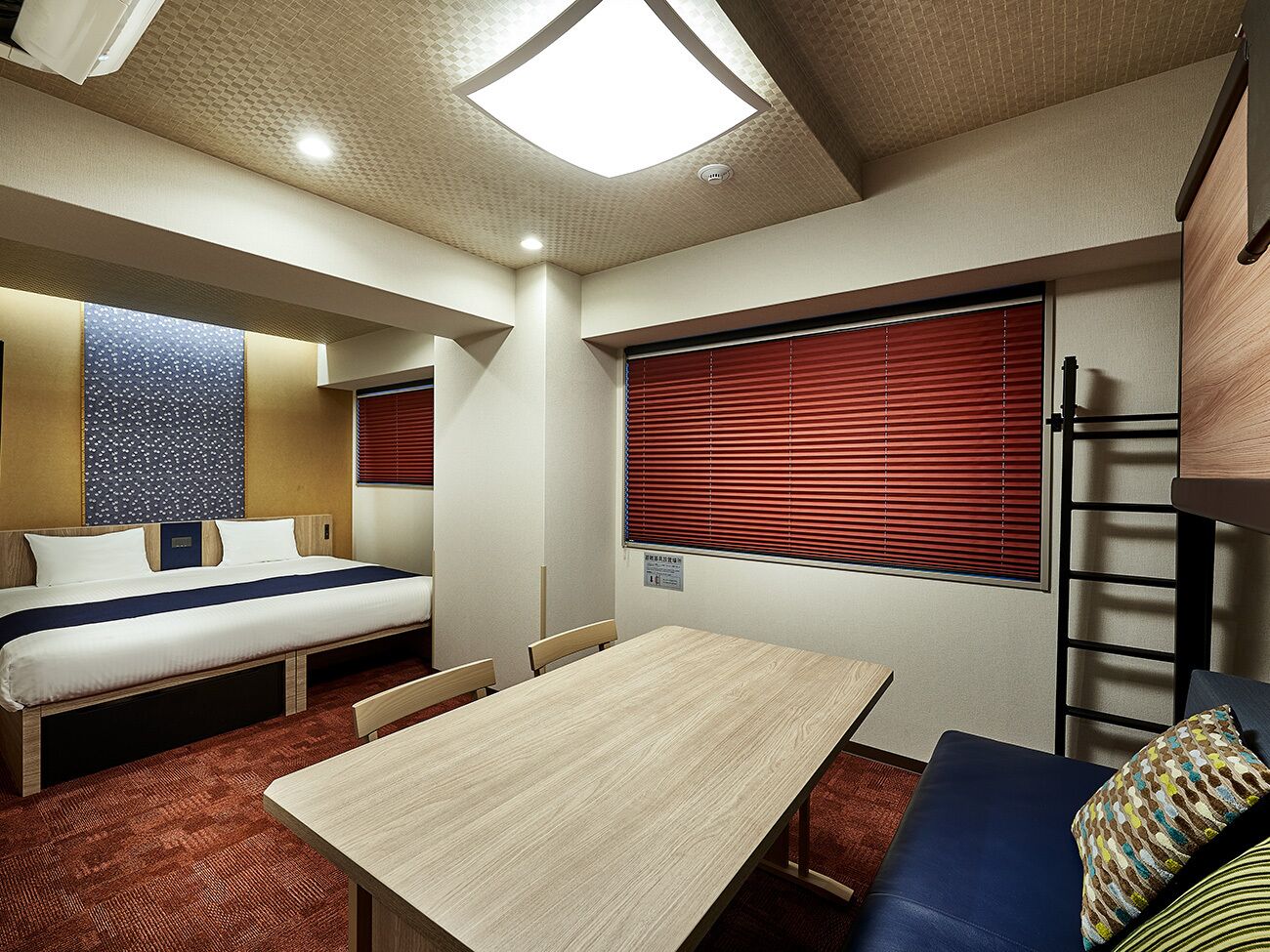 A compact hotel room with a bed, a small table, a sofa, and a window with blinds.