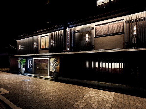 A modern hotel exterior at night with illuminated signage, a potted plant, and a paved entrance.