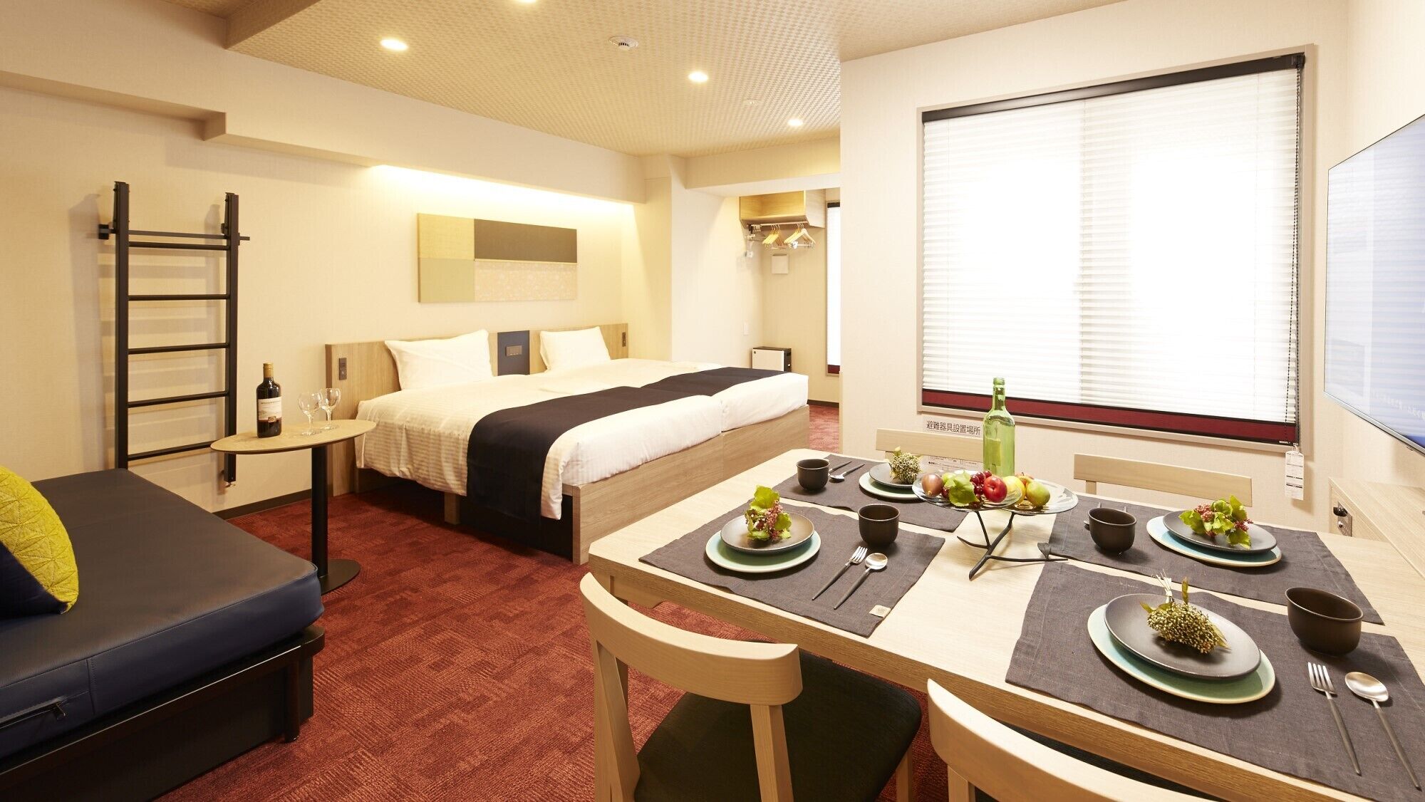 Habitación de hotel con cama, una mesa de comedor con platos y cubiertos, una mesita con una botella y vasos, un sofá y una ventana con persianas.