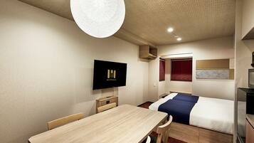 A compact hotel room with a wooden dining table, a single bed, a TV mounted on the wall, and a small kitchenette.