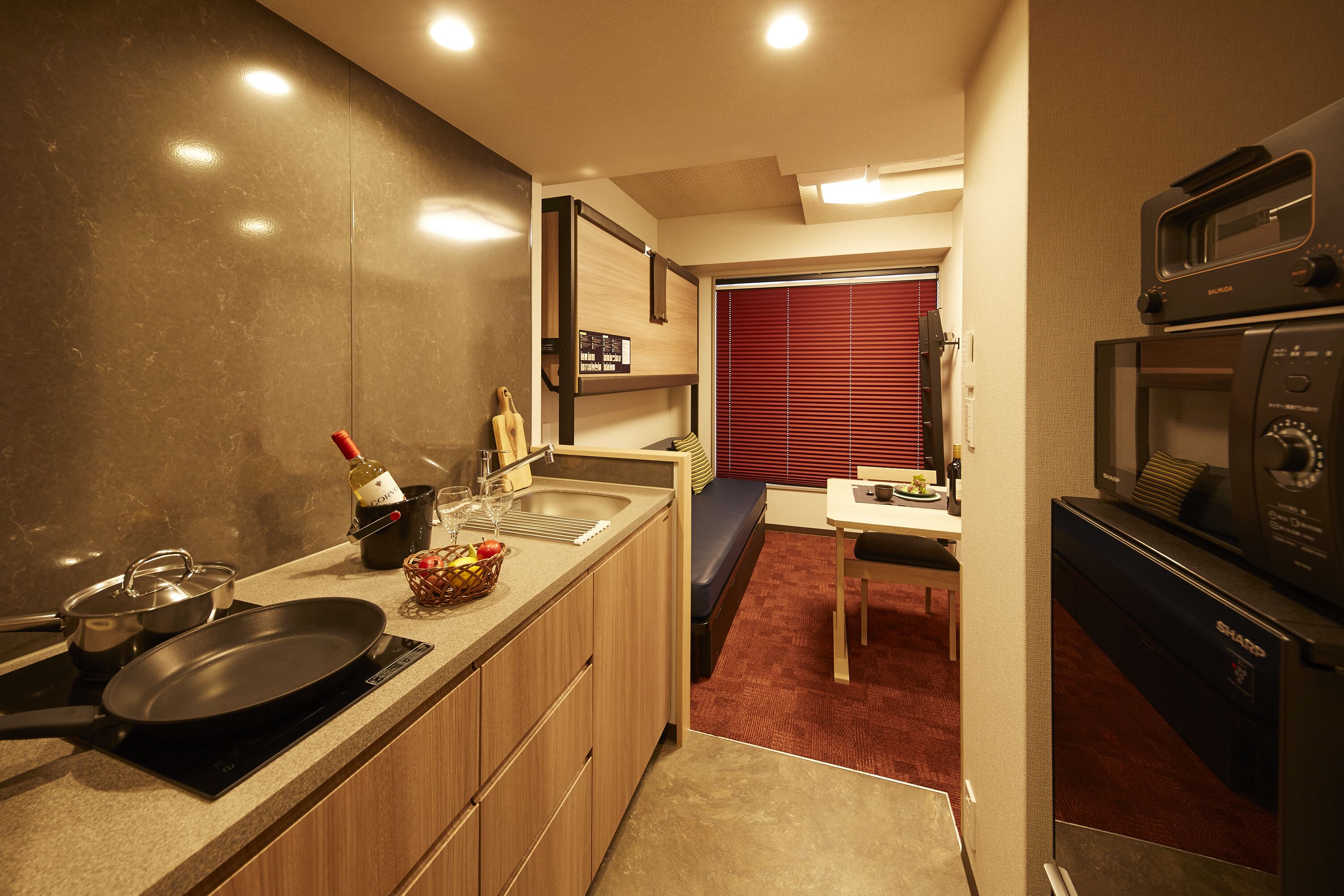 A compact kitchen with a stove, sink, and microwave. There is a dining area with a bench and a window with blinds.