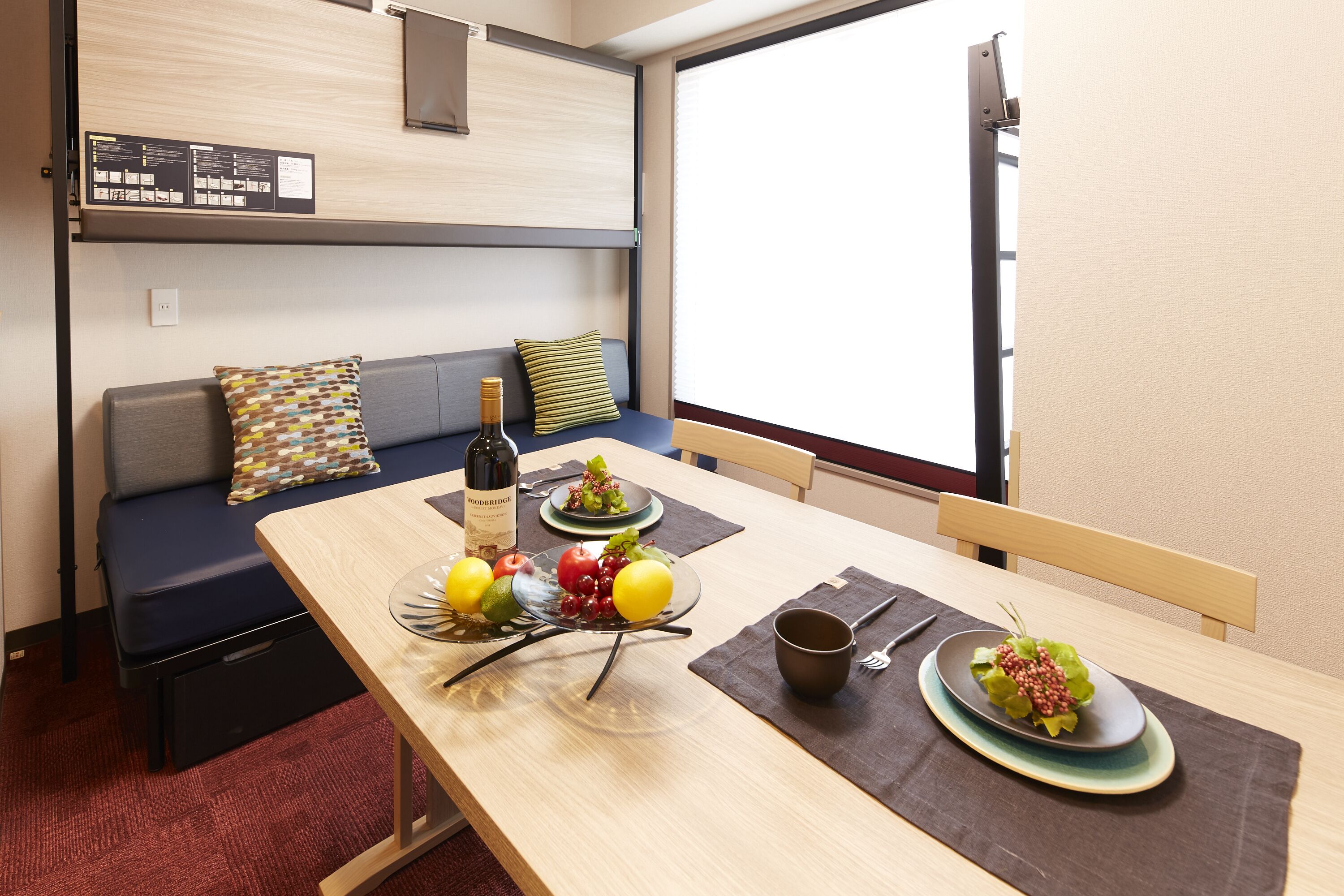 A compact room with a dining table set for two, a sofa with cushions, a window with blinds, and a wall-mounted information board.