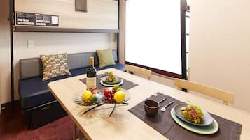 A compact room with a dining table set for two, a sofa with cushions, a window with blinds, and a wall-mounted information board.