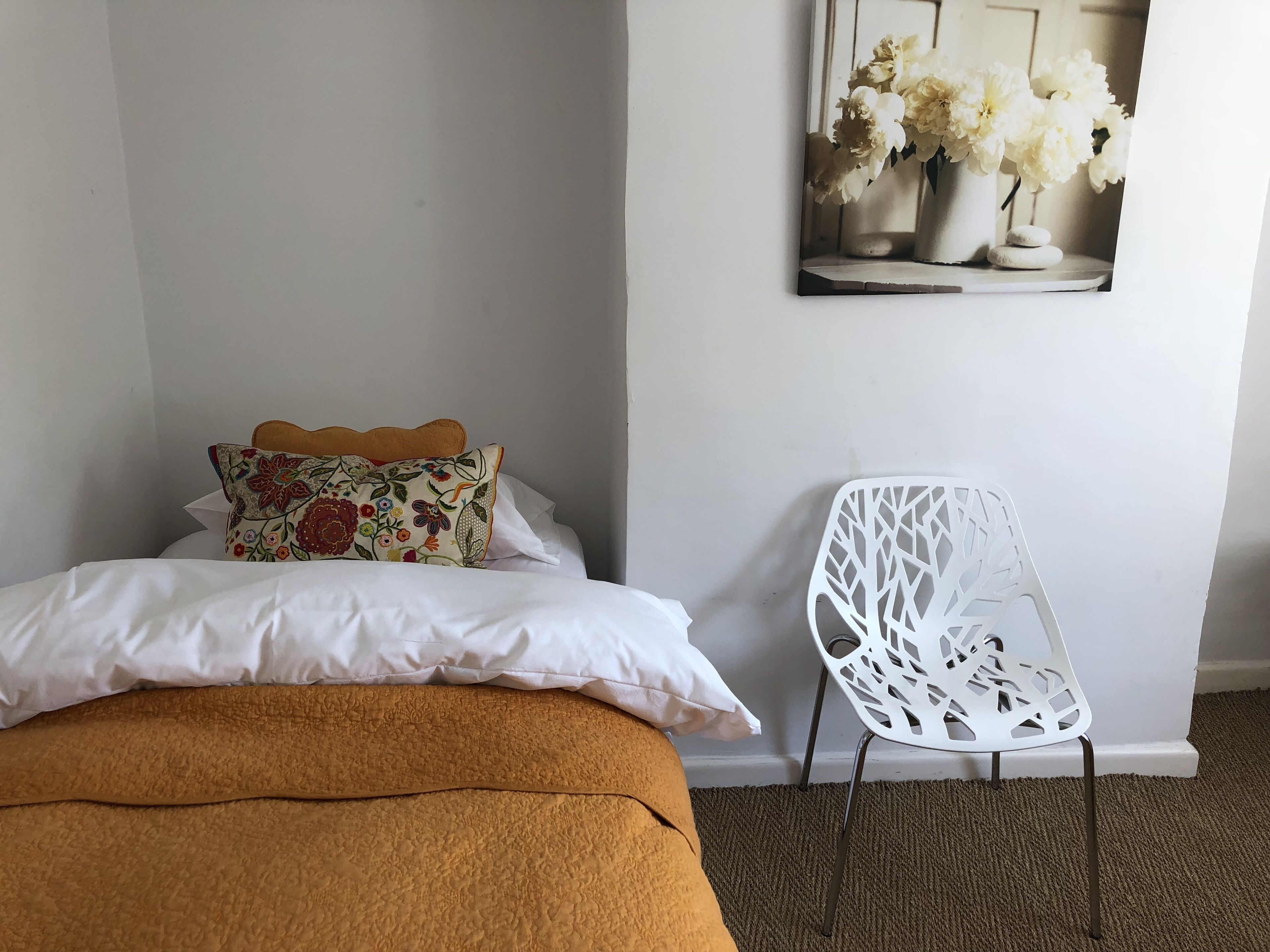 A bedroom with a bed, a decorative pillow, a white chair, and a wall-mounted picture of flowers.