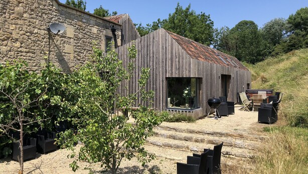 A rustic stone building with a wooden extension, outdoor seating, and a gravel path.