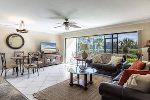 A living room with a television, a ceiling fan, a dining area with chairs and a table, and a large window with a view of palm trees.