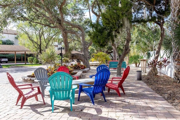 Outdoor seating with Adirondack chairs and a table, surrounded by trees and a paved walkway.