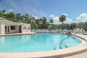 A swimming pool with lounge chairs and umbrellas, surrounded by palm trees and a building with a door.
