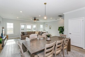 A spacious living room with a large wooden dining table, a ceiling fan, and a view of the outdoors through large windows.