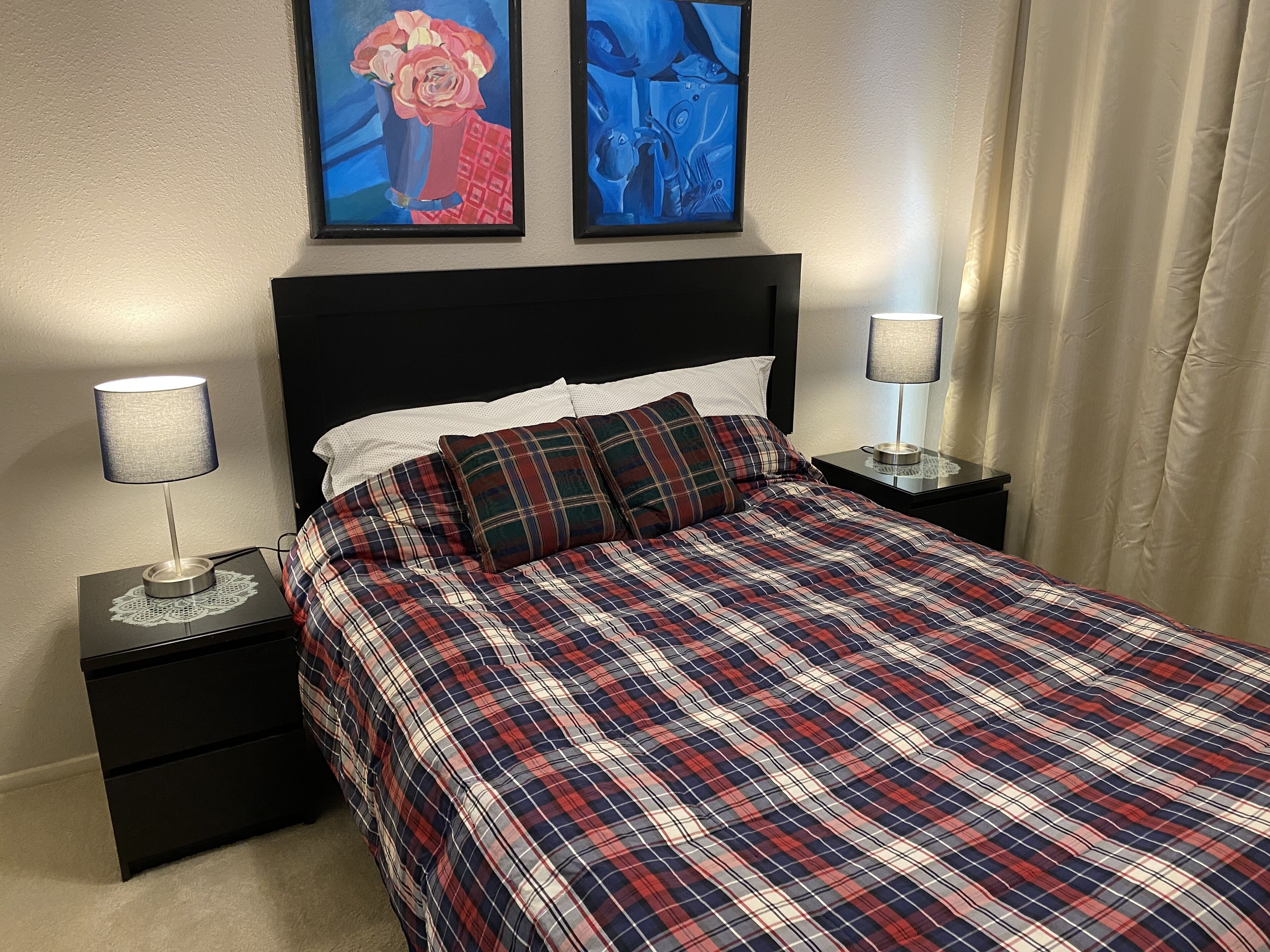 A bedroom with a plaid bedspread, two bedside tables with lamps, a black headboard, and two framed artworks on the wall.