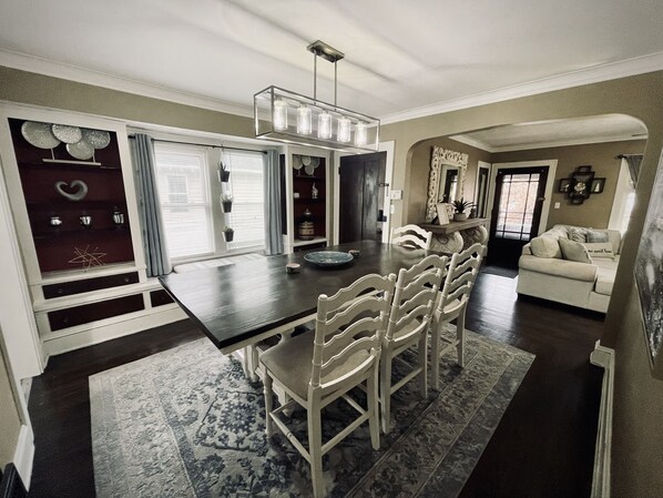 Un coin repas avec une table en bois foncé, des chaises blanches et un lustre. On aperçoit un canapé et un miroir décoratif à l’arrière-plan.