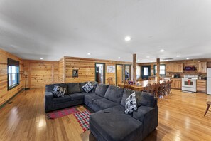 A modern log cabin interior with a large gray sectional sofa, wooden floors, a kitchen with white appliances, and a dining area.