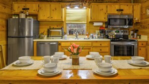 A wooden cabin kitchen with a stainless steel refrigerator, microwave, stove, and sink. There is a dining table set with white cups and saucers, and a vase with pink flowers. - 🌲Easy Paved Access🔥Fireplace❤️Great Privacy🏞️Across River🎇NearDowntown🙂 (Blue Ridge)
