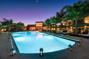 Une piscine à bord incurvé, entourée de chaises longues et de palmiers, avec des bâtiments en arrière-plan.