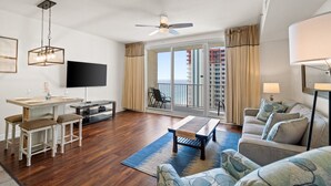 A spacious living room with a wooden floor, a dining area with a table and chairs, a sofa set, a flat-screen TV, and a balcony with a view of the city. - Summer Deals!! Gulf Front, Beach Access + Parking! 17th Floor! Winter $1800/mo! (Panama City Beach)