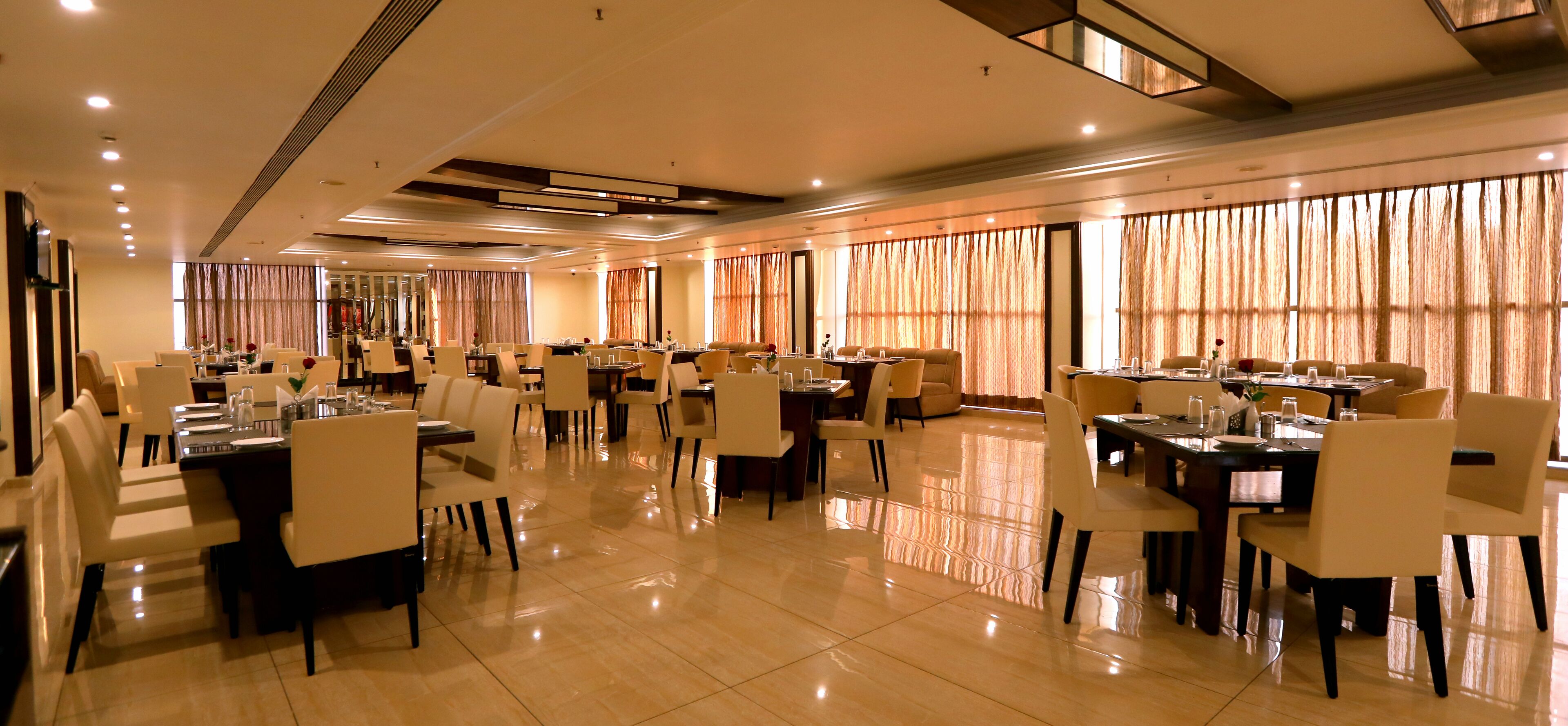 Ampla área de jantar de hotel, com várias mesas e cadeiras, grandes janelas com cortinas e piso polido.