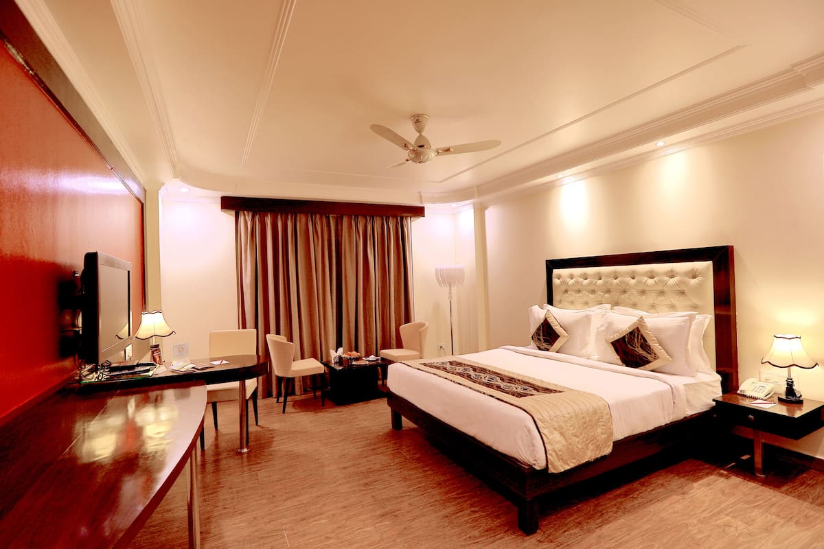 A hotel room with a large bed, a desk, two chairs, a ceiling fan, and a nightstand with a lamp.