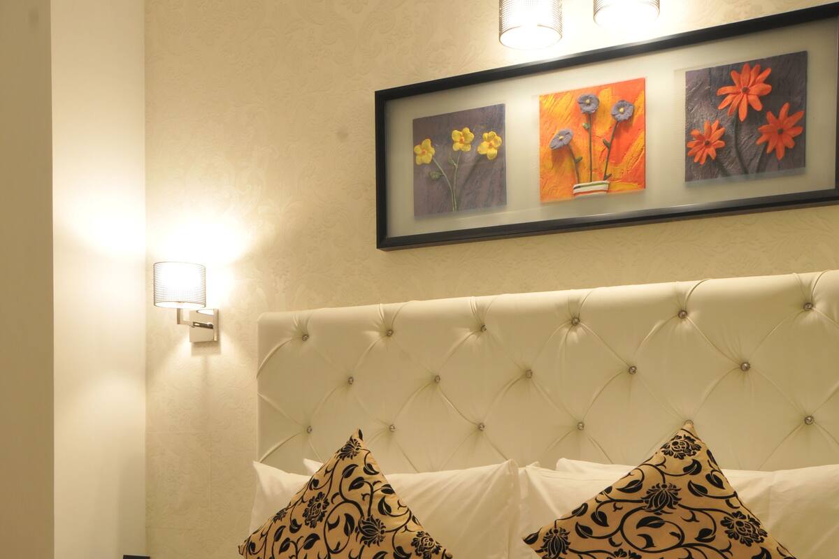 A hotel room with a tufted headboard, floral-patterned pillows, a neatly made bed, and a framed floral artwork above the bed.