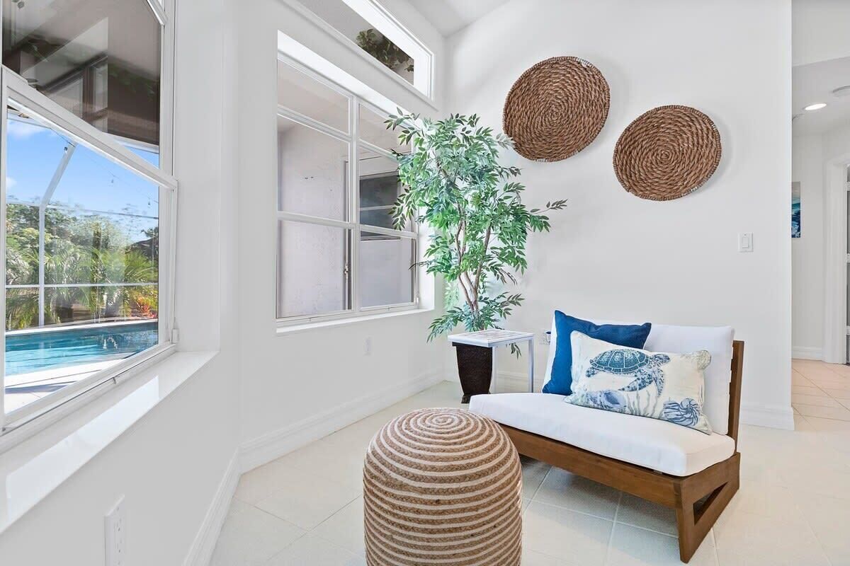 A bright room with a large window, a wooden sofa with a blue pillow, a woven ottoman, and a potted plant.